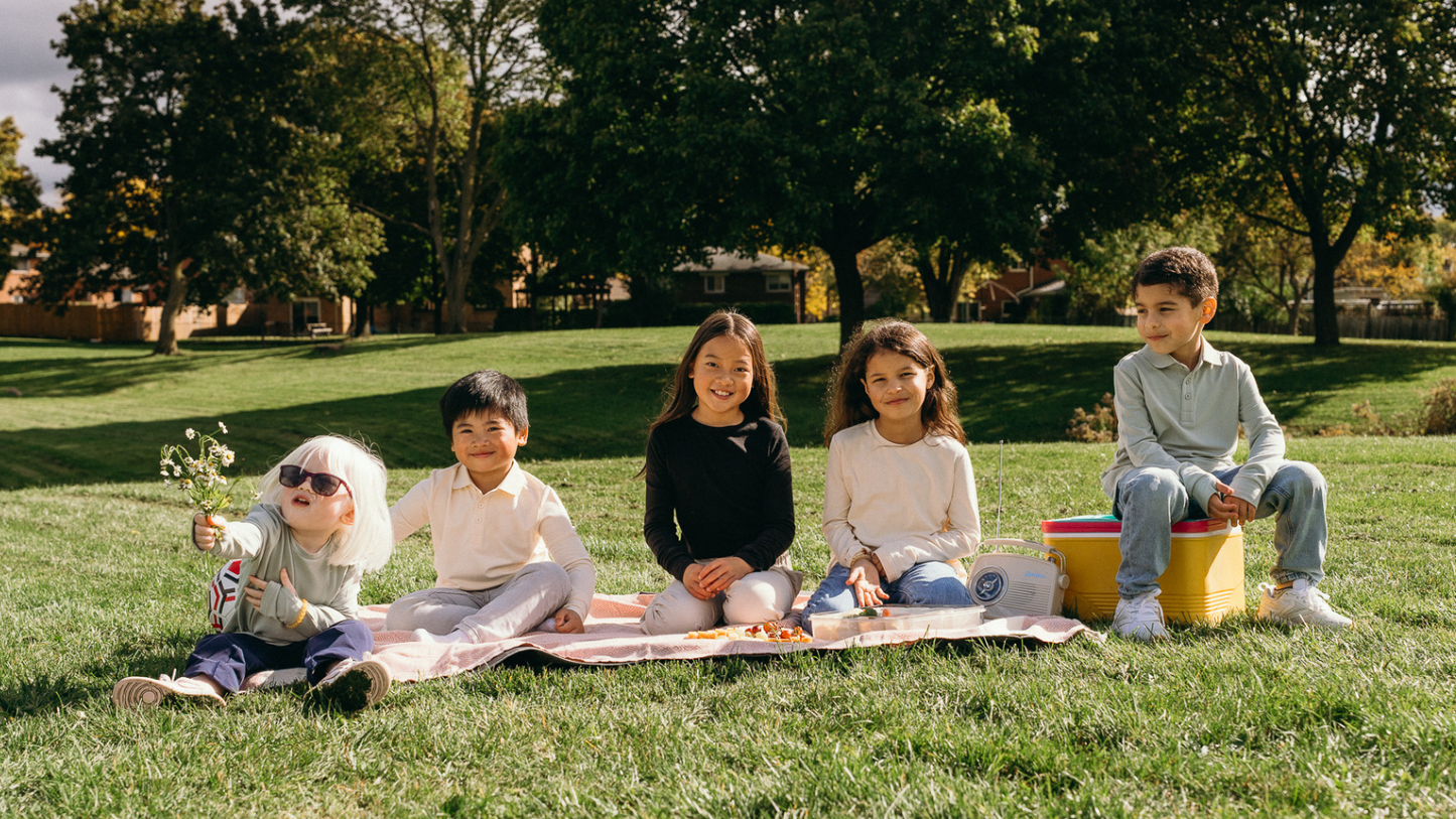Five children sitting on a picnic blanket in a sunny park wearing Ember & Ray UPF 50+ clothing